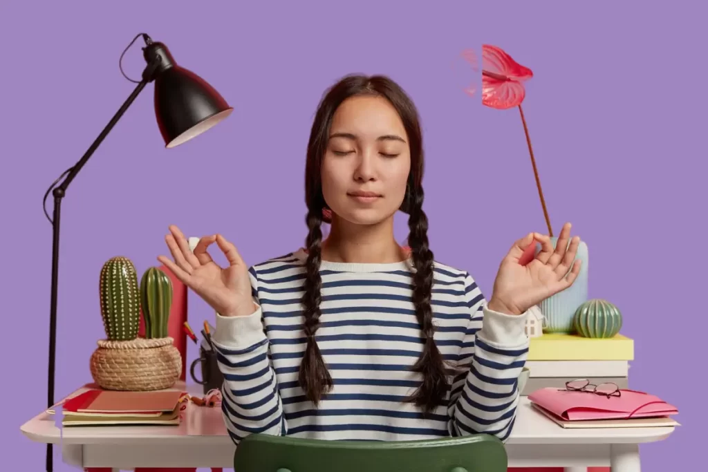 Fotos, mulher asiática relaxada medita no local de trabalho, senta se em pose zen contra a mesa com flores, abajur, blocos de notas, usa um macacão casual listrado, tenta relaxar depois do trabalho