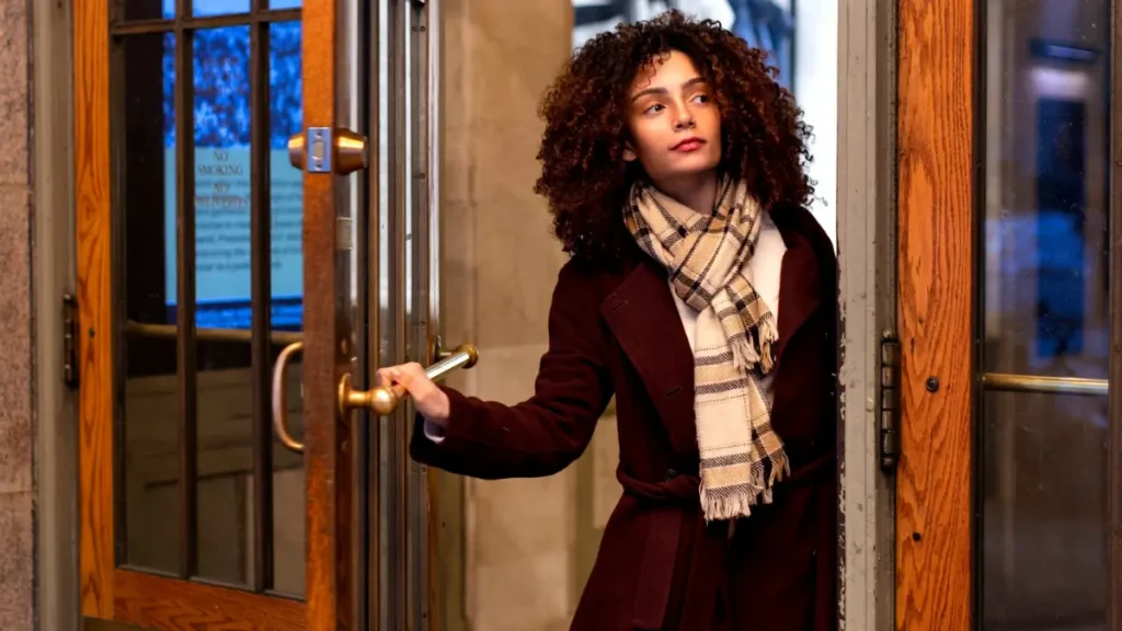 Mulher adulta brasileira, cerca de 35 anos, aparência natural e realista, cabelos cacheados castanho-escuros, parada diante de uma porta aberta que leva para um ambiente iluminado, expressão pensativa e levemente insegura, uma das mãos próxima à maçaneta como se estivesse prestes a entrar, simbolizando medo de começar e autossabotagem.