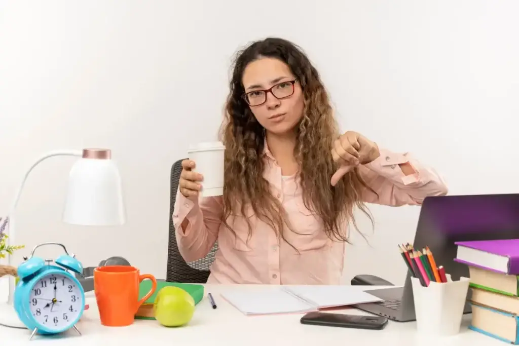 Imagem de stockking no Freepik Jovem bonita insatisfeita usando óculos, sentada na mesa com as ferramentas de Rotina Produtiva, segurando a xícara de café e mostrando o polegar para baixo