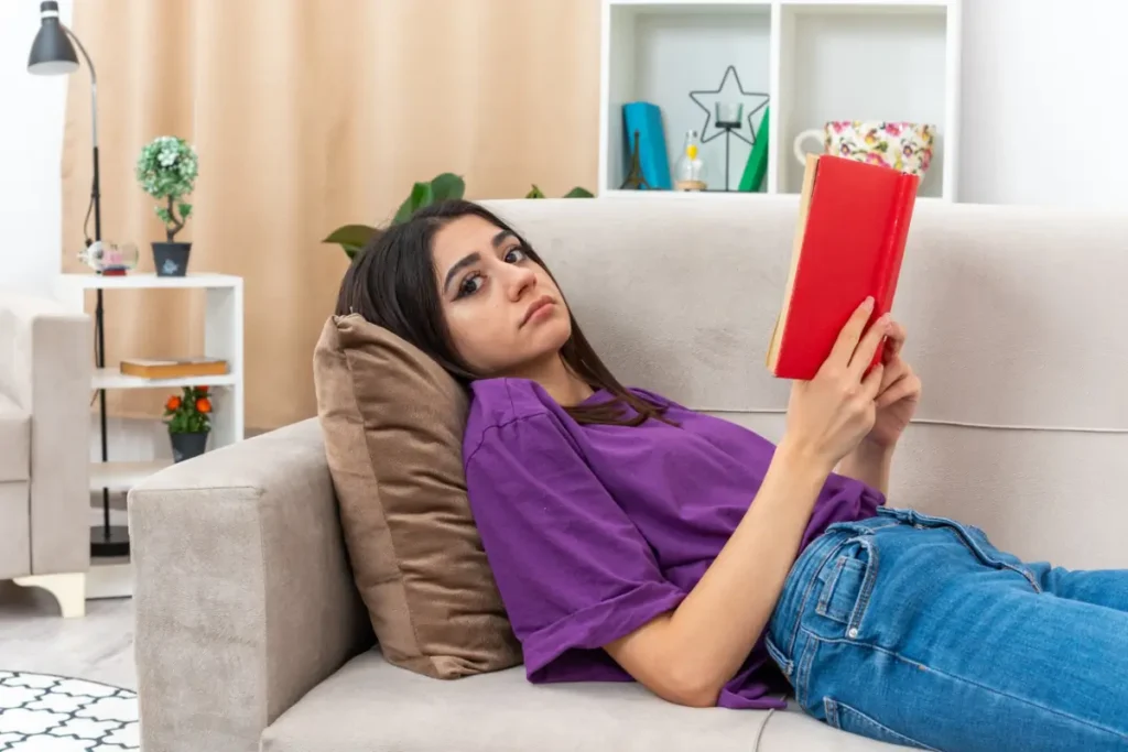 _Jovem com roupas casuais, com um livro, com uma expressão cansada, deitada em um sofá na sala de estar iluminada. Imagem de stockki
