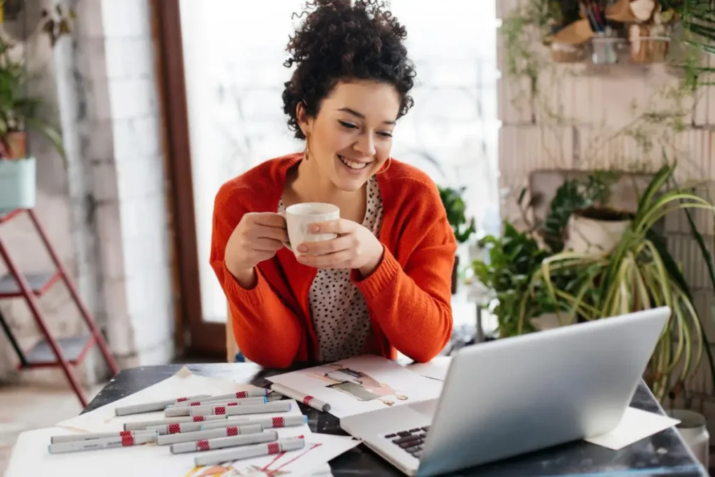 Jovem linda, mulher sorridente com cabelos cacheados escuros, sentada à mesa segurando o copo na mão, na mesa com agenda, celular, lista de tarefas e trabalhando alegremente no lapt